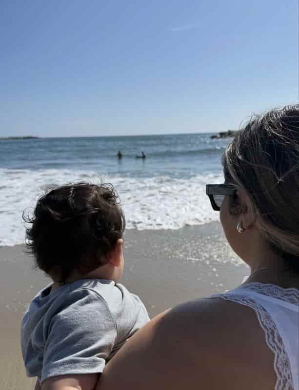 Mom holding her baby son at the beach as he watches the ocean waves for the first time
