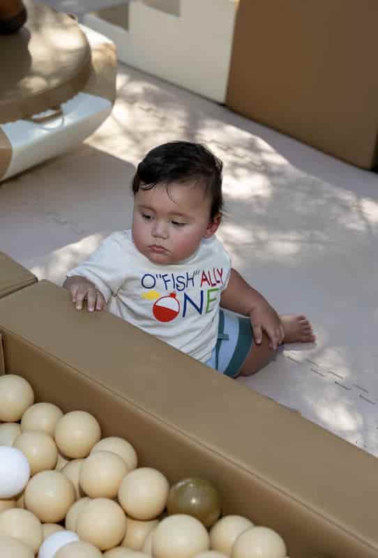 One-year-old boy in an “O-fish-ally One” first birthday shirt sitting in a ball pit